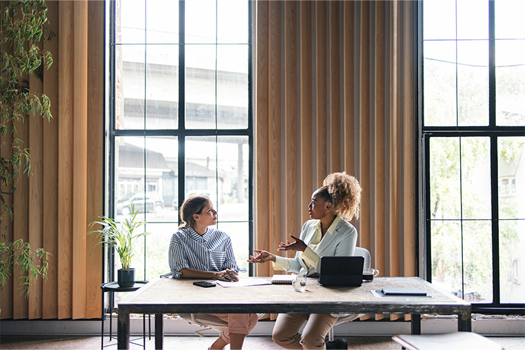 Two women catching up at a desk in a modern, professional setting 
