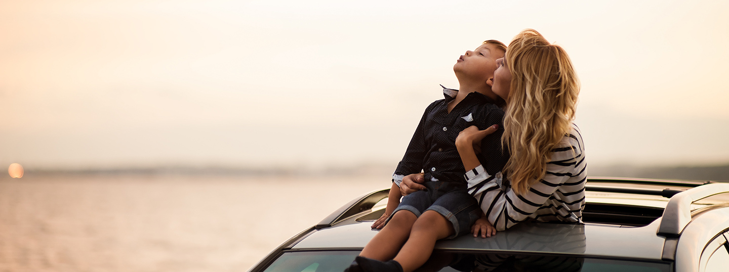 Woman hugging her son, who's sitting on the car's roof, through the sunroof