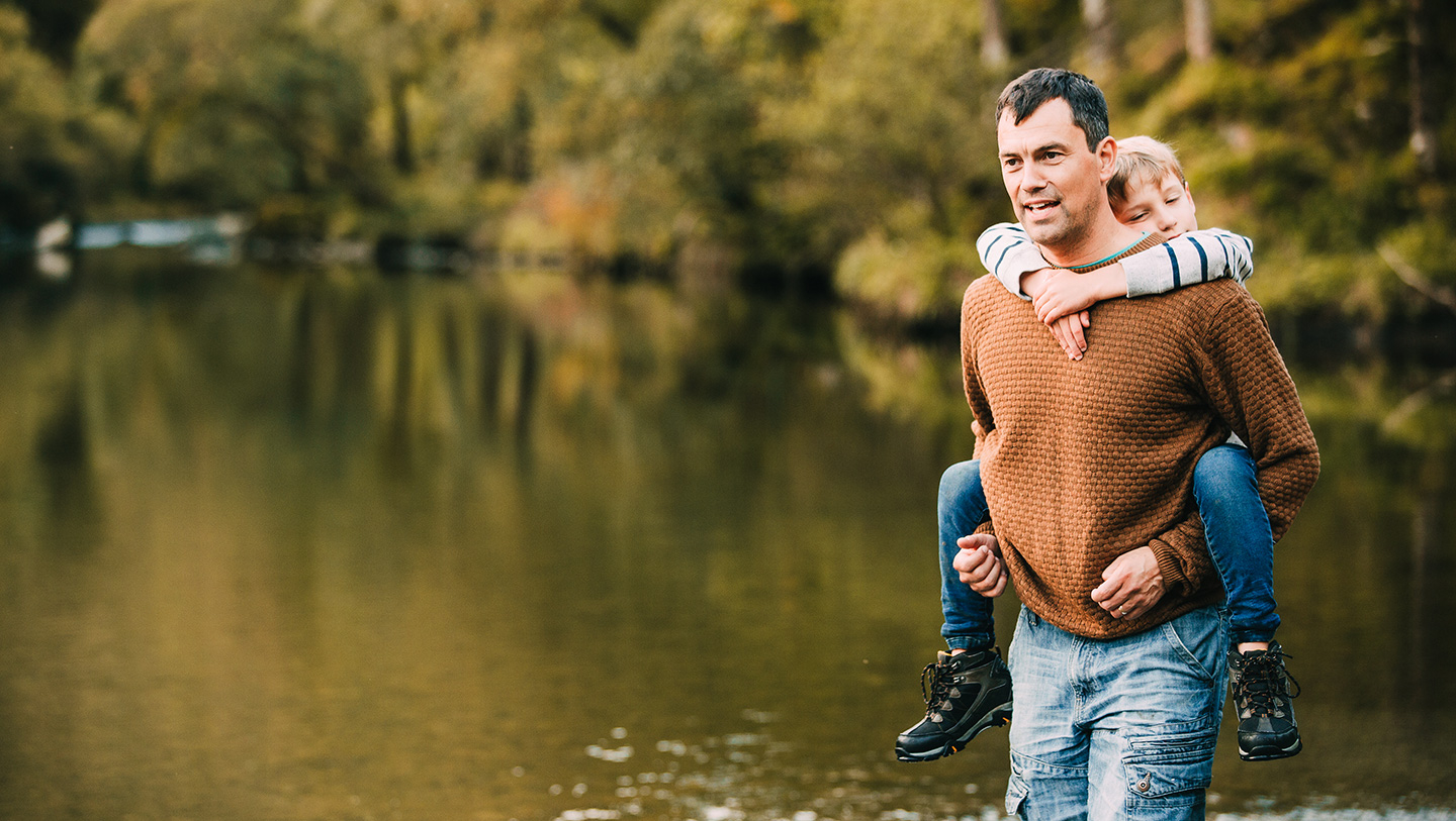 Middle aged man carrying his son in front of a lake in the woods