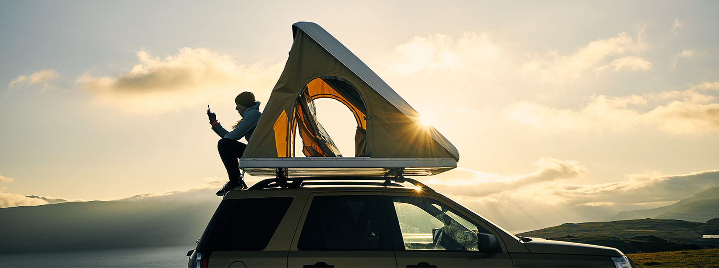Person sitting on a car roof with a roof-mounted tent, enjoying the scenery at sunset