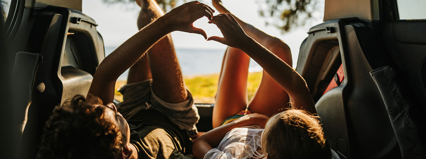 Two children reclining in the back of a vehicle at sunset, touching hands to form a heart shape