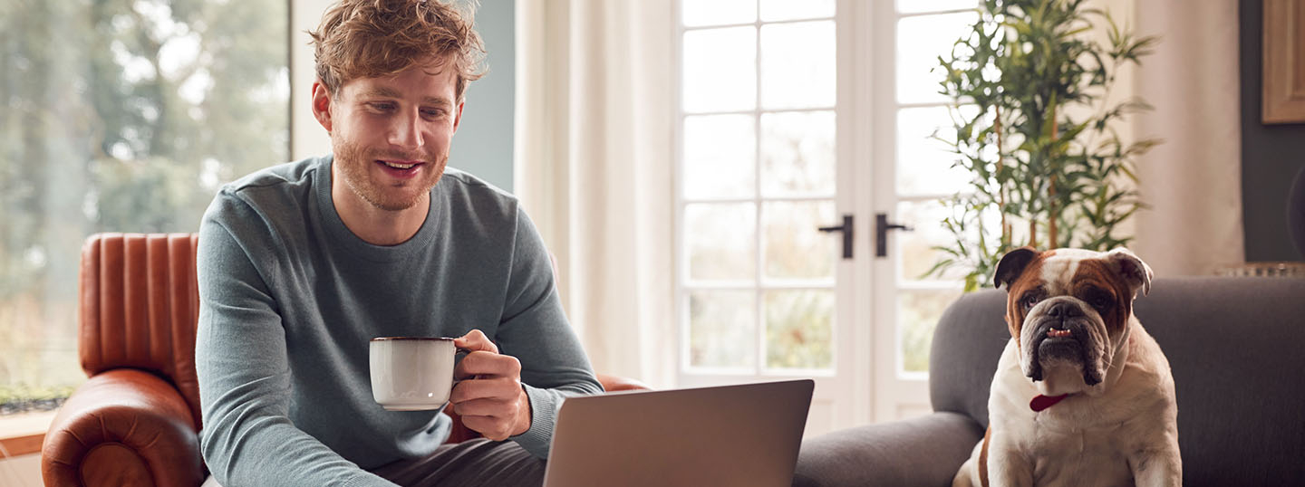 Image of a young man having a cup of coffee and happily checking his laptop, with a dog sat in the next chair along
