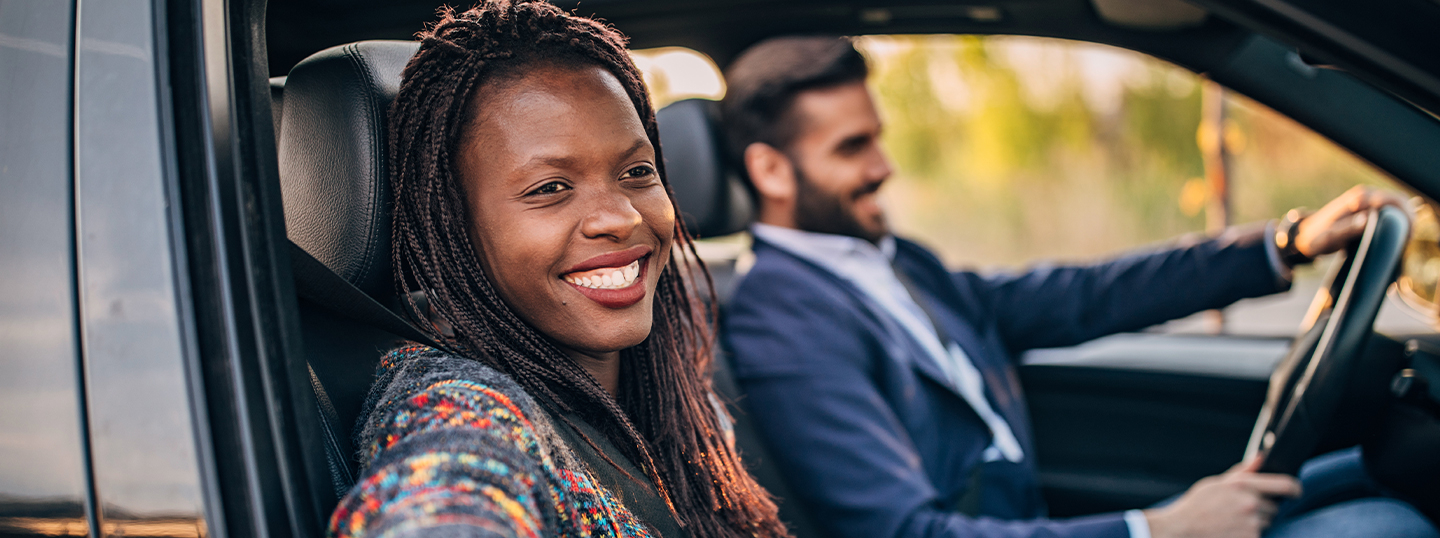 A smiling Black woman with braids looking out the window of the passenger seat with her partner driving in the background 