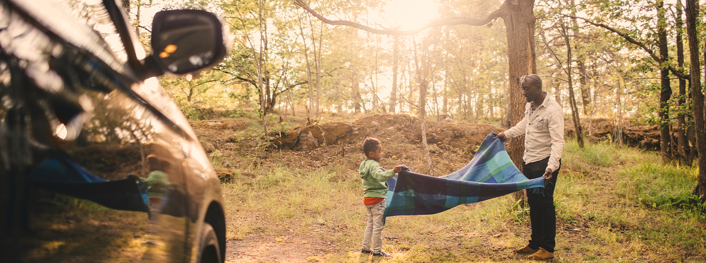 A father and son laying out a picnic blanket in a woodland setting, with their new car in the foreground 