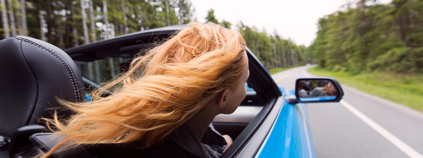 Image of a blonde person in a convertible, hair flying freely, happily driving and concentrating on the road ahead