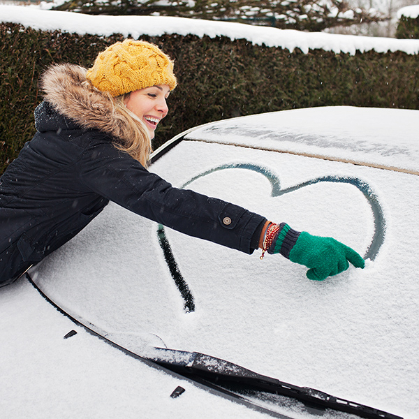 Woman smiling and writing a heart into the snow on her car's windscreen