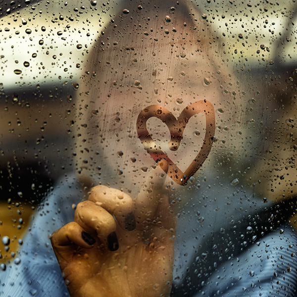 A person smiling and drawing a heart in the condensation on a car window