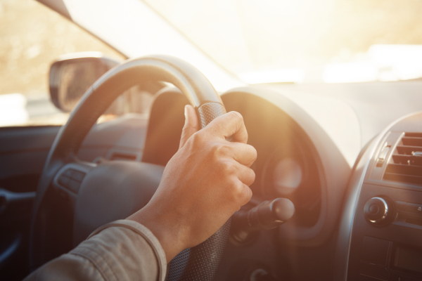 A person's hand on the steering wheel close up with sun-glare reflecting off the windscreen