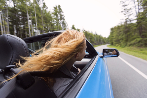 a long-haired person looking in the mirror of her blue convertible while driving through woodland