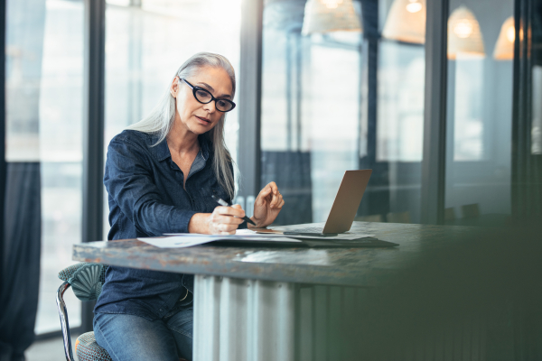 A grey-haired businesswoman reviewing documents carefully on her laptop 